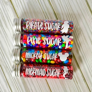 May include: Four clear glass tubes filled with colorful sprinkles and candies. Each tube has a silver cap and a white label with text and a silhouette: "Pirate Sugar", "Pixie Sugar", "Mickey Sugar", and "Mermaid Sugar".