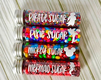 May include: Four clear glass tubes filled with colorful sprinkles and candies. Each tube has a silver cap and a white label with text and a silhouette: "Pirate Sugar", "Pixie Sugar", "Mickey Sugar", and "Mermaid Sugar".