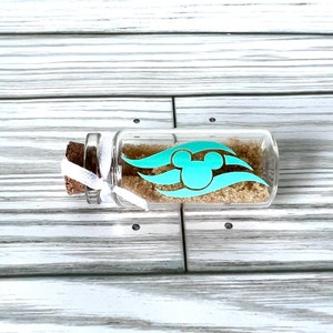 May include: A small, clear glass bottle filled with brown granules, sealed with a cork and tied with a white ribbon. A turquoise graphic of Mickey Mouse and stylized waves adorns the bottle. The bottle is displayed on a light-colored wooden surface.