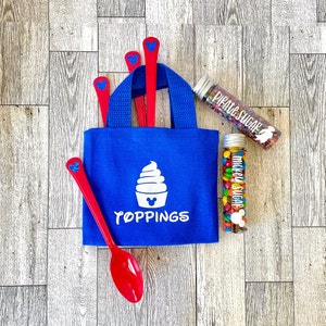 May include: A blue tote bag with the word "TOPPINGS" and a white ice cream cone graphic. Red spoons with a blue Mickey Mouse head design are in the bag. Two clear tubes contain colorful sprinkles and candies, labeled "PIRATE SUGAR" and "MICKEY SUGAR."