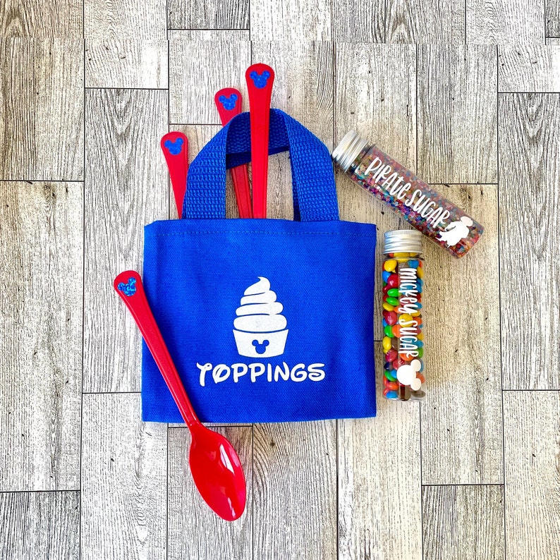 May include: A blue tote bag with the word "TOPPINGS" and a white ice cream cone graphic. Red spoons with a blue Mickey Mouse head design are in the bag. Two clear tubes contain colorful sprinkles and candies, labeled "PIRATE SUGAR" and "MICKEY SUGAR."