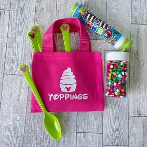 May include: A vibrant pink tote bag with the word "TOPPINGS" and a cupcake graphic. The bag contains green spoons with a glittery mouse ear design. A tube of M&M's Minis and a jar of star-shaped sprinkles are also present.