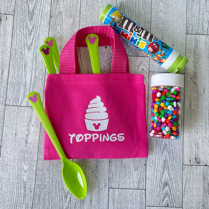 May include: A vibrant pink tote bag with the word "TOPPINGS" and a cupcake graphic. The bag contains green spoons with a glittery mouse ear design. A tube of M&M's Minis and a jar of star-shaped sprinkles are also present.
