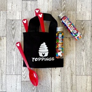May include: A black tote bag with the word "TOPPINGS" and a white ice cream design. Red spoons with Mickey Mouse head cutouts are in the bag. Two clear tubes filled with colourful sweets and the words "Mickey Sugar" and "Pixie Sugar" are also present.
