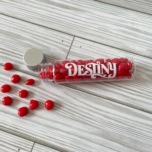 May include: A clear plastic tube filled with red candies, with the word "DESTINY" in white script. The tube is lying on its side, with some candies spilled out. The background is a light gray wooden surface.