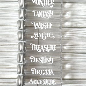 May include: Eight clear glass vials with silver-colored screw-on lids. Each vial has a white cursive word: Wonder, Fantasy, Wish, Magic, Treasure, Destiny, Dream, and Adventure. The vials are arranged vertically against a light wood-grain background.