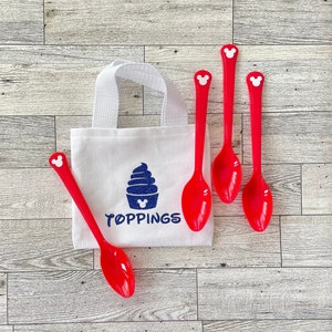 May include: A white canvas tote bag with a blue ice cream graphic and the word "TOPPINGS" in blue. Four red plastic spoons with Mickey Mouse head details are arranged around the bag. The background is a light wood grain.