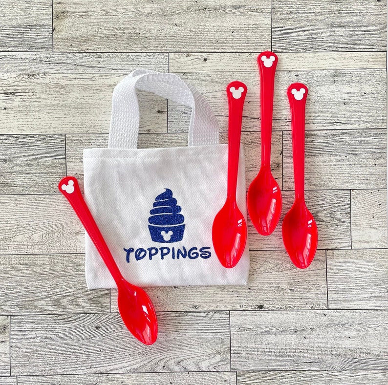 May include: A white canvas tote bag with a blue ice cream graphic and the word "TOPPINGS" in blue. Four red plastic spoons with Mickey Mouse head details are arranged around the bag. The background is a light wood grain.