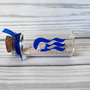 May include: A small, clear glass bottle filled with sand and sealed with a cork. A blue ribbon is tied around the neck of the bottle. A blue graphic of stylized waves is on the bottle. The bottle is lying on a light wood surface.