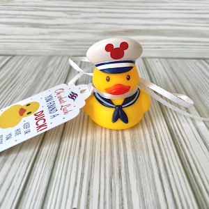 May include: A yellow rubber duck wearing a sailor hat with a red Mickey Mouse emblem and a navy blue necktie. A white tag with the text "YOU FOUND A DUCK!" is attached with a white ribbon. The duck is on a light wood-grain surface.