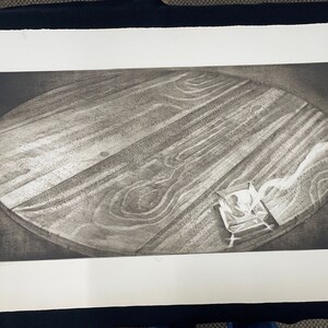 May include: A black and white print of a round wooden table with a small object on the surface. The table is made of wood with a grain pattern. The object is a small, white, rectangular box with a design on the top.