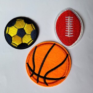 Peut inclure: Trois patchs brodés thermocollants représentant un ballon de football, un ballon de basket-ball et un ballon de rugby. Le ballon de football est jaune et noir, le ballon de basket-ball est orange et noir, et le ballon de rugby est rouge et blanc.