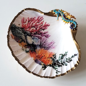 Puede incluir: Un plato decorativo en forma de concha blanca con borde dorado. El interior presenta un diseño de arrecife de coral colorido con tonos rojos, naranjas y morados. Un caballito de mar azul y dorado adorna el borde. Ideal para decoración costera.