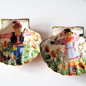 Puede incluir: Dos conchas decorativas con ribetes dorados, cada una con un espantapájaros pintado en un entorno de jardín. Un espantapájaros lleva una camisa de cuadros rojos y negros y pantalones vaqueros azules, mientras que el otro lleva un vestido rosa y blanco. Ambos espantapájaros tienen un sombrero de paja y una cara amigable.