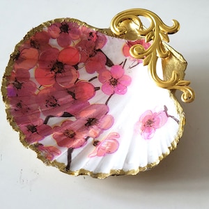 May include: A decorative seashell dish with a floral design. The white shell is adorned with pink and red flowers and a gold-painted edge. A gold-colored, ornate metal accent is attached to the shell.