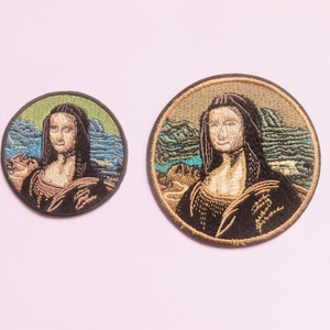 May include: Two embroidered patches featuring the Mona Lisa, one larger than the other. The patches are circular and have a brown background with a green and blue landscape in the background. The Mona Lisa is depicted in a black dress with a gold necklace.