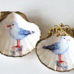 Op de afbeelding: Twee decoratieve schelpen met een gouden rand en een geschilderd zeemeeuwontwerp. Eén schelp heeft een gouden kroon met blauwe accenten. De zeemeeuwen zitten op houten palen met touwdetails.
