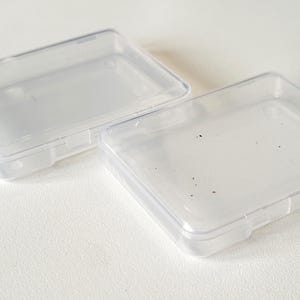 Può includere: Due contenitori di plastica trasparenti rettangolari con coperchi incernierati. Un coperchio è aperto, l'altro è chiuso. I contenitori sono su una superficie bianca. La plastica è traslucida e i contenitori sembrano vuoti.