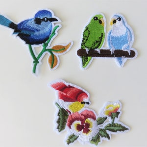 May include: Three embroidered iron-on patches featuring birds. One patch shows a blue bird perched on a green branch. Another patch shows two birds, one green and one blue, perched on a brown branch. The third patch shows a red and yellow bird with a white flower.