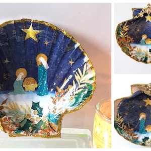 Può includere: Una conchiglia blu e dorata con una scena della Natività dipinta su di essa. La scena include Maria, Giuseppe e il bambino Gesù. La conchiglia è decorata con glitter dorato e stelle.