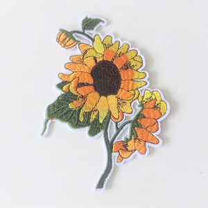 Peut inclure: Patch brodée thermocollante représentant un tournesol jaune avec un cœur brun et des feuilles vertes. Le tournesol est entouré de petites fleurs jaunes.