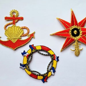Op de afbeelding: Drie geborduurde strijkbare patches: een rode anker met een gouden schelp, een rode ster met een gouden middelpunt en een gele reddingsboei met rode en blauwe accenten.