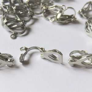 Peut inclure: Clips d'oreille en métal argenté. Les clips sont conçus pour maintenir les boucles d'oreilles en place. Il y a plusieurs clips éparpillés sur une surface blanche.