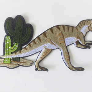 Peut inclure: Un patch brodé thermocollant représentant un dinosaure brun et blanc avec des cactus verts.