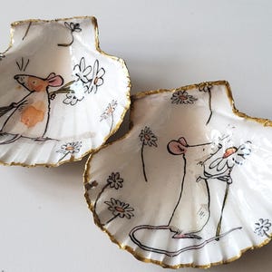 Könnte beinhalten: Zwei dekorative Muschelschalen mit einem skurrilen Design. Jede Schale zeigt eine handgemalte Illustration einer Maus, die Blumen hält, mit einem goldfarbenen Rand. Der Hintergrund ist weiß mit verstreuten Gänseblümchen-Akzenten.