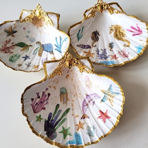 May include: Three white seashell-shaped dishes with gold trim. Each dish is decorated with a different underwater scene, including starfish, seahorses, a dolphin, a jellyfish, and a turtle.