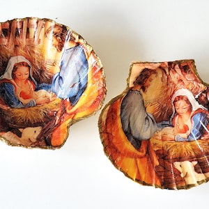 Può includere: Due ornamenti a conchiglia raffigurano una scena religiosa con figure in blu, bianco e oro. L'opera d'arte raffigura una donna che tiene in braccio un bambino, con un uomo in una veste blu. Le conchiglie hanno un bordo dorato.