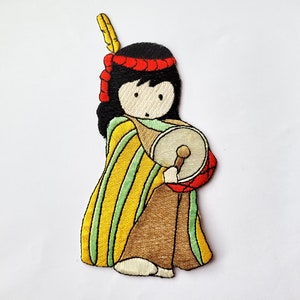 May include: Embroidered patch of a cartoon Native American person wearing a yellow and green striped robe, a red headband, and holding a small drum.