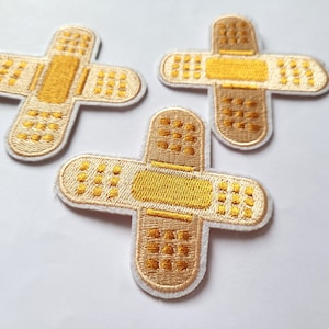 Peut inclure: Trois patchs en tissu blanc en forme de pansements avec un centre jaune et une bordure blanche. Les patchs sont brodés d'un motif de petits carrés.