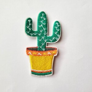 Peut inclure: Écusson brodé représentant un cactus vert avec des détails blancs, planté dans un pot jaune avec des accents orange et verts. Conçu pour être appliqué sur des vêtements ou des accessoires.