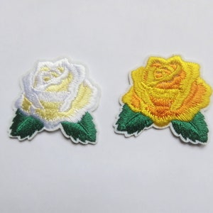 Peut inclure: Deux patchs brodés à repasser en forme de roses. Une rose est blanche avec des accents jaunes et l'autre est jaune avec des accents orange. Les deux roses ont des feuilles vertes.