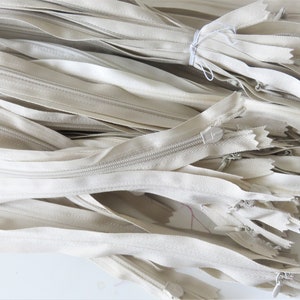 May include: A pile of off-white zipper closures. The zippers are all the same length and have a white pull tab.