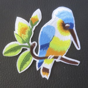 Peut inclure: Patch thermocollant brodé représentant un oiseau bleu, jaune et orange perché sur une branche avec des feuilles vertes.