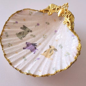 May include: A decorative seashell trinket dish with a gold-painted rim and accents. The interior features a white background with a floral pattern and illustrations of three cartoon bunnies in green, purple, and yellow.