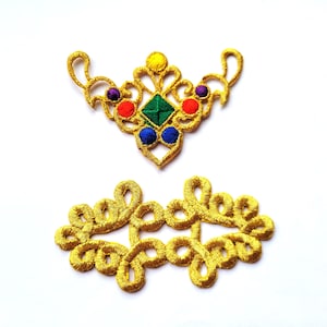 Peut inclure: Deux embellissements brodés dorés avec des perles colorées. L'embellissement du haut est en forme de cœur avec un motif de perles vertes, bleues, rouges et jaunes. L'embellissement du bas est un motif plus complexe avec des boucles et des volutes.