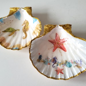 Könnte beinhalten: Zwei weiße Muschelschalen mit goldenen Verzierungen. Eine zeigt einen Seestern, Seepferdchen und Korallen. Die andere hat eine Meeresschildkröte und ein Seepferdchen.