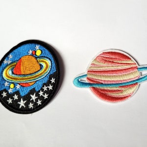 Peut inclure: Deux patchs brodés thermocollants représentant des planètes avec des anneaux. Un patch est bleu avec une planète jaune et orange et des étoiles blanches. L'autre patch est rose avec une planète blanche et rose et un anneau bleu.