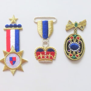 Könnte beinhalten: Drei gestickte Aufnäher mit einem Stern, einer Krone und einem runden Design mit einem blauen Juwel. Der Stern-Aufnäher hat ein rot-weiß-blaues Band. Der Kronen-Aufnäher ist rot und gold mit einem blauen Zentrum. Der runde Aufnäher ist schwarz mit goldenen, grünen und roten Akzenten.