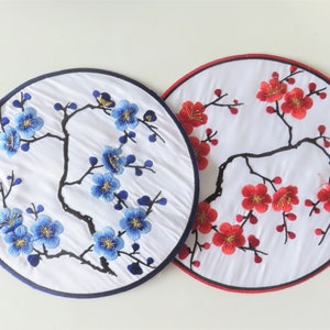 Peut inclure: Deux panneaux ronds en tissu brodé avec un fond blanc et une branche noire avec des fleurs bleues et rouges. Les fleurs ont des accents dorés.
