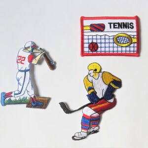 May include: Three embroidered iron-on patches featuring sports themes. The first patch shows a baseball player in a red and blue uniform swinging a bat. The second patch shows a hockey player in a yellow, red, and blue uniform holding a hockey stick. The third patch is a rectangular patch with a tennis net and the word "TENNIS" in red.