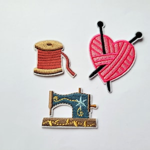 May include: Three embroidered iron-on patches: a spool of red thread, a pink heart with two knitting needles, and a blue sewing machine.