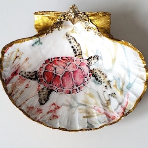 Può includere: Una conchiglia bianca con un bordo dorato, decorata con una tartaruga marina rosa dipinta e una barriera corallina. La conchiglia ha un manico dorato decorato.