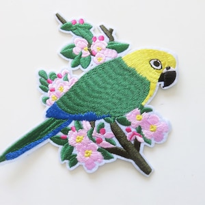 Peut inclure: Patch brodée thermocollante représentant un perroquet vert et jaune perché sur une branche avec des fleurs roses.