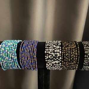 May include: A display of six beaded bracelets in various colors. The bracelets are arranged on a black stand. Colors include shades of blue, white, gold, black, and silver. Each bracelet appears to be approximately 1 inch wide.