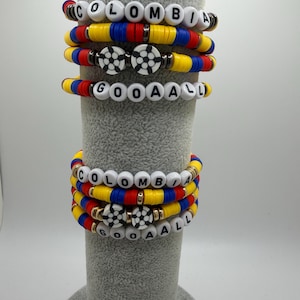 May include: A collection of vibrant bracelets, displaying the word "COLOMBIA" and "GOOAAAL" in white beads. The bracelets incorporate the yellow, blue, and red of the Colombian flag, with soccer ball designs.