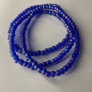 K&ouml;nnte beinhalten: Eine blaue Perlenkette mit schillernden Perlen. Die Kette besteht aus mehreren Perlenstr&auml;ngen und ist auf einer wei&szlig;en Oberfl&auml;che aufgerollt.